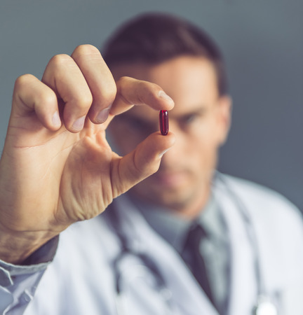 Handsome medical doctor in white coat is holding a pill, on gray background. Hand with medicine in focusの写真素材