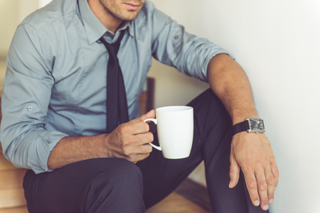 Cropped image of handsome businessman in classic suit holding a cup of coffee while sitting on the stairs at homeの写真素材