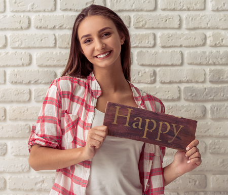 Attractive young girl in casual clothes is holding a wooden plate "happy", looking at camera and smiling, on white brick wall backgroundの写真素材