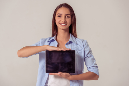 Attractive young girl in casual clothes is showing a digital tablet, looking at camera and smiling, on gray backgroundの写真素材
