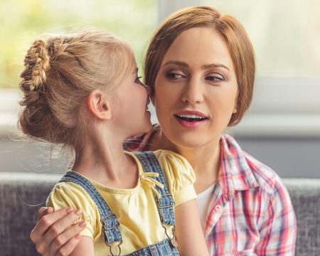 Cute little girl is whispering something in her beautiful mother's ear. Both are sitting on couch at homeの写真素材