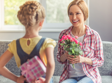 Beautiful mother is holding flowers, looking at her cute little daughter and smiling while sitting on couch at home. Girl is holding a gift box behind her backの写真素材