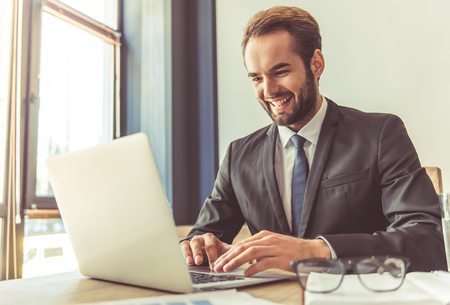 Attractive businessman in formal suit is smiling while working with a laptop in his officeの写真素材