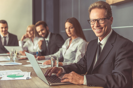 Business people in formal wear are discussing affairs at the conference. Handsome man in the foreground is using a laptop, looking at camera and smilingの写真素材