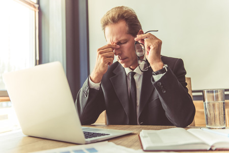 Handsome tired businessman in formal suit is holding eyeglasses and massaging his nose bridge while working with a laptop in his officeの写真素材