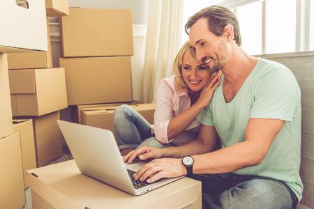Beautiful mature couple in casual clothes is using a laptop, hugging and smiling while sitting among...