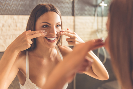 Beautiful young woman in white lingerie is applying cream on her face and smiling while looking into the mirror in bathroomの写真素材