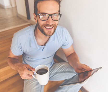 Handsome young man in eyeglasses is using a digital tablet, holding a cup of coffee, looking at camera and smiling while sitting on stairs at homeの写真素材