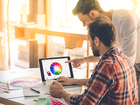 Handsome designers in casual clothes are using laptop and discussing colors while working in studioの写真素材