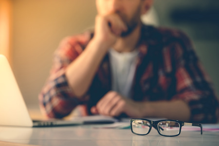 Eyeglasses on table, in the background tired designer is leaning on his hand while working in his...