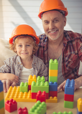Handsome grandpa and grandson in helmets are looking at camera and smiling while playing with construction set at homeの写真素材