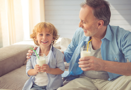 Handsome grandpa and grandson are drinking milk and smiling while spending time together at home. Little boy is looking at cameraの写真素材