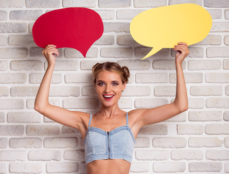 Beautiful stylish girl in jeans top and white shorts is holding speech bubbles, looking at camera and smiling, on white brick wall backgroundの写真素材