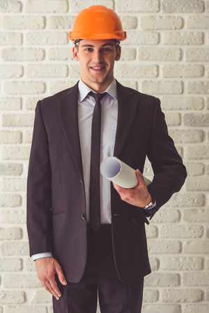 Handsome young businessman in suit and protective helmet is holding rolled paper, looking at camera and smiling, on a white brick wall backgroundの写真素材