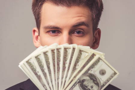 Portrait of handsome young businessman in suit holding money and looking at camera, on a gray backgroundの写真素材