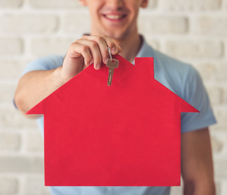 Cropped image of handsome muscular guy in blue t-shirt holding a paper house and a key and smiling, on a white brick wall backgroundの写真素材