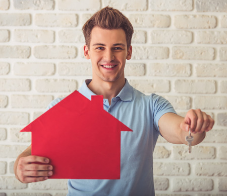 Handsome muscular guy in blue t-shirt is holding a paper house and a key, looking at camera and smiling, on a white brick wall backgroundの写真素材