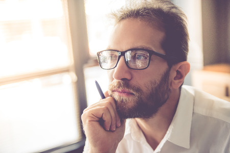 Portrait of handsome pensive young businessman in eyeglasses holding a pen, looking away and thinkingの写真素材