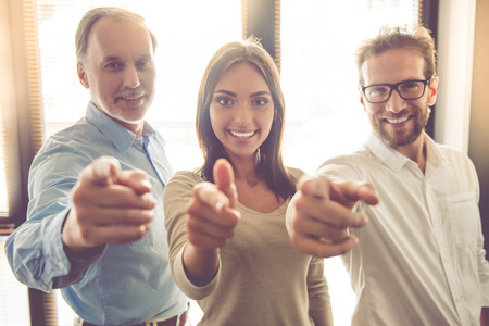 Beautiful business team in smart casual wear are smiling, looking and pointing at camera while standing in their officeの写真素材