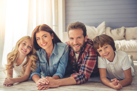 Beautiful young parents and their children are looking at camera and smiling while lying together on the floor at homeの写真素材