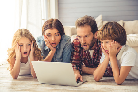 Beautiful young parents and their children are using a laptop and showing surprise while lying together on the floor at homeの写真素材
