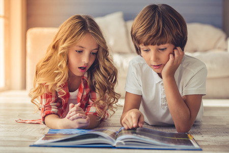 Pretty little girl and boy are reading book and showing surprise while lying on the floor at homeの写真素材