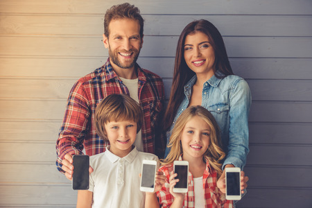Beautiful young parents and their children are holding smartphones, looking at camera and smiling,...