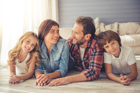 Beautiful young parents and their children are looking at camera and smiling while lying together on the floor at homeの写真素材