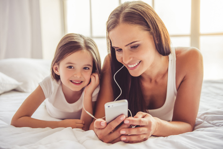 Beautiful young woman and her charming little daughter in earphones are listening to music using a smart phone and smiling while lying in bed at homeの写真素材