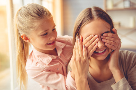 Portrait of beautiful young mother and her little daughter smiling while sitting on sofa at home. Girl is covering mom's eyesの写真素材