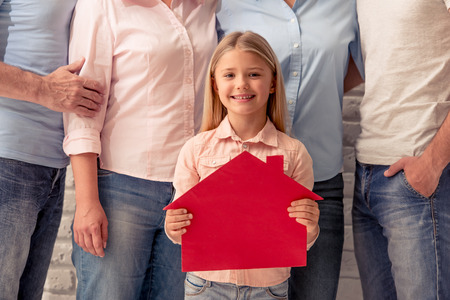 Cute little girl is holding a paper house, looking at camera and smiling, her beautiful parents and grandparents are standing behindの写真素材