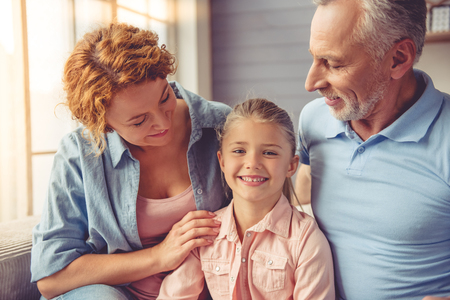 Happy mature couple and their little granddaughter are hugging, talking and smiling while sitting on sofa at homeの写真素材
