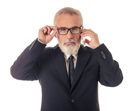 Handsome bearded mature businessman in classic suit and eyeglasses is talking on the mobile phone and looking at camera, isolated on whiteの写真素材