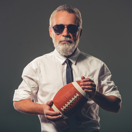 Handsome bearded mature businessman in classic shirt and glasses is holding a football ball and looking at camera, on a gray backgroundの写真素材