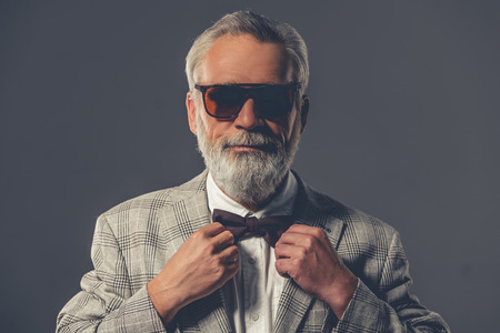 Handsome bearded mature gentleman in formal suit and glasses is adjusting his bow tie, looking at camera and smiling, on a gray backgroundの写真素材