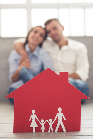 A red paper house, in the background beautiful young couple is hugging and smiling while sitting on the floor at homeの写真素材
