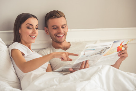 Beautiful young couple is reading a newspaper, talking and smiling while lying in bed in the morningの写真素材