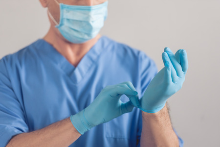 Cropped image of handsome mature surgeon in blue medical wear and mask putting on medical gloves, on gray backgroundの写真素材