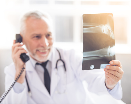 Handsome mature medical doctor in white coat is talking on the phone, examining x-ray picture and smiling while working in his officeの写真素材