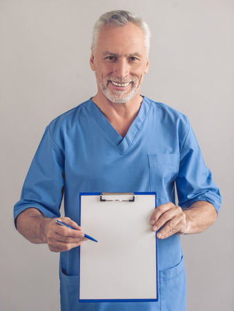 Handsome mature surgeon in blue medical wear is holding a folder, looking at camera and smiling, on gray backgroundの写真素材