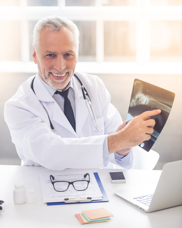 Handsome mature medical doctor in white coat is pointing on x-ray picture, looking at camera and smiling while working in his officeの写真素材