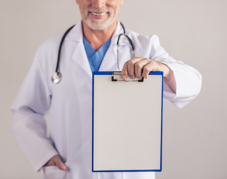 Cropped image of handsome mature medical doctor in white medical coat holding a folder and smiling, on gray backgroundの写真素材