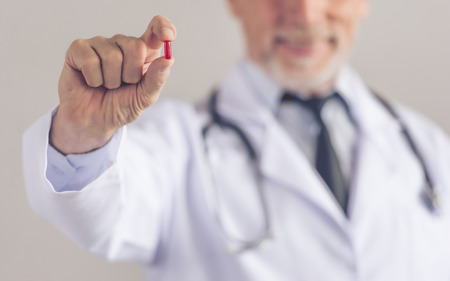 Cropped image of handsome mature medical doctor in white coat holding a pill and smiling, on gray background. Medicine in focusの写真素材