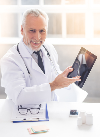 Handsome mature medical doctor in white coat is pointing on digital tablet, looking at camera and smiling while working in his officeの写真素材
