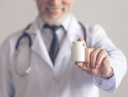 Cropped image of handsome mature medical doctor in white coat holding a bottle of medicine and smiling, on gray background. Medicine in focusの写真素材
