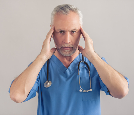 Handsome tired mature medical doctor in blue medical wear is holding hands on his temples and looking at camera, on gray backgroundの写真素材