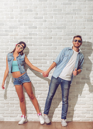 Full length portrait of stylish teenage couple in jeans clothes and sn glasses holding hands, looking at camera and smiling, on white brick wall backgroundの写真素材