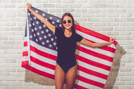 Stylish young girl in black bodysuit and sun glasses is holding American flag, looking at camera and smiling, on white brick backgroundの写真素材
