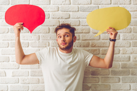 Handsome guy in casual clothes is holding speech bubbles, looking at camera and showing dismay, on white brick wall backgroundの写真素材