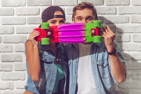 Stylish teenage couple in jean clothes and caps is holding skateboard, hiding behind it and looking at camera, on white brick wall backgroundの写真素材